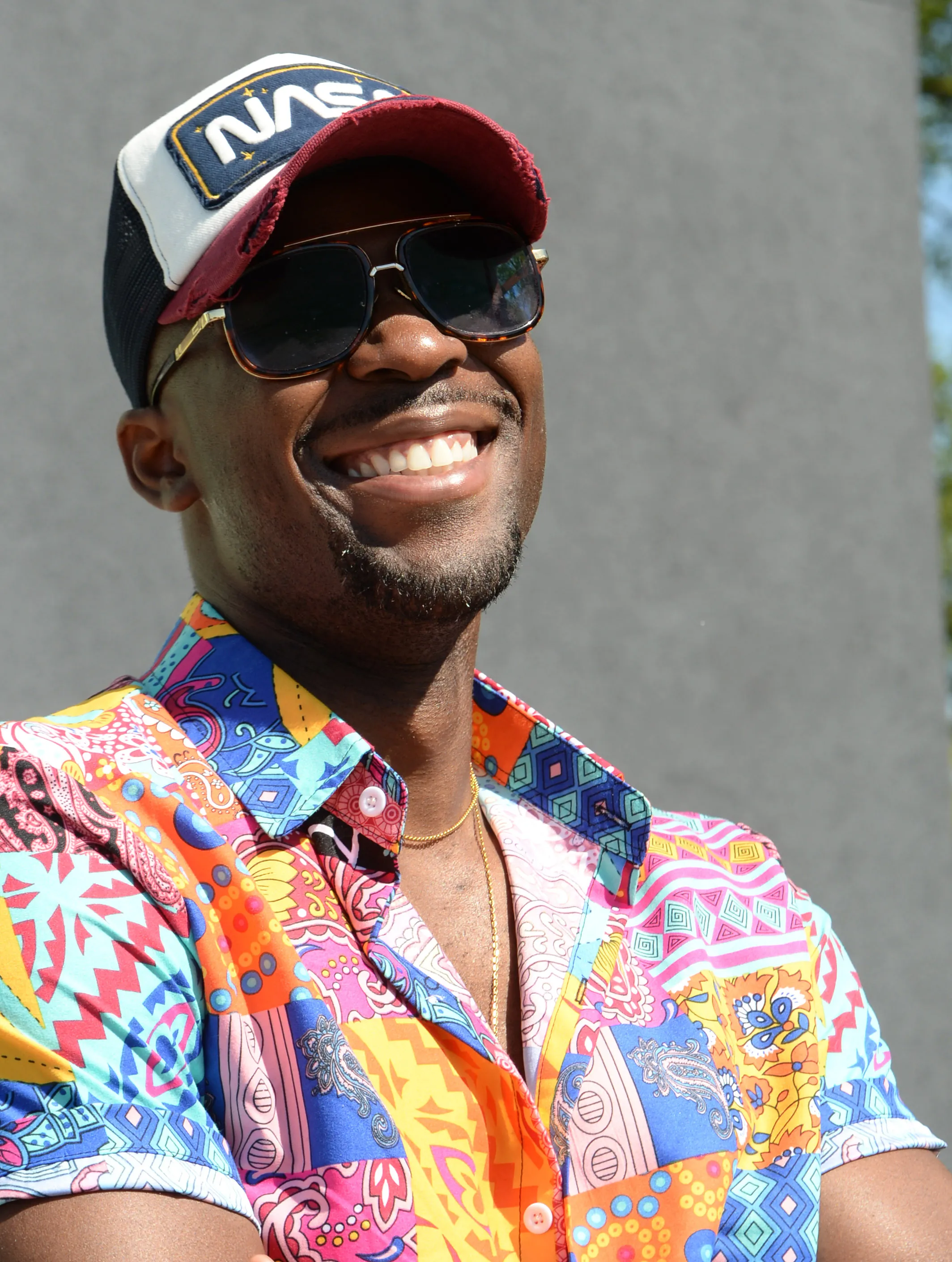 A portrait of a man who is smiling and wearing a NASA baseball cap, sunglasses and a colorful print shirt.