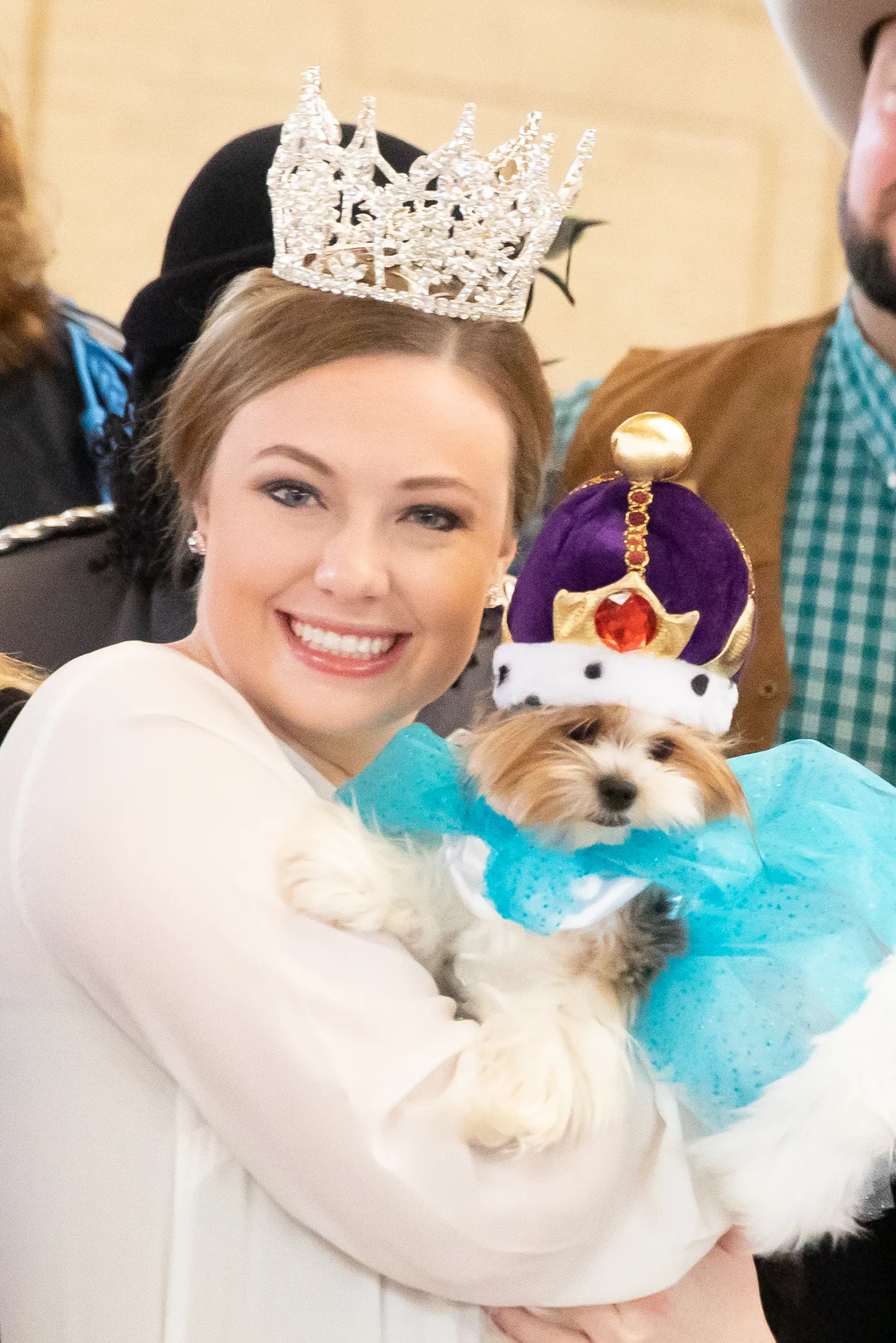 A woman wearing a crown smiles as she poses holding a tiny dog who is also wearing a crown.