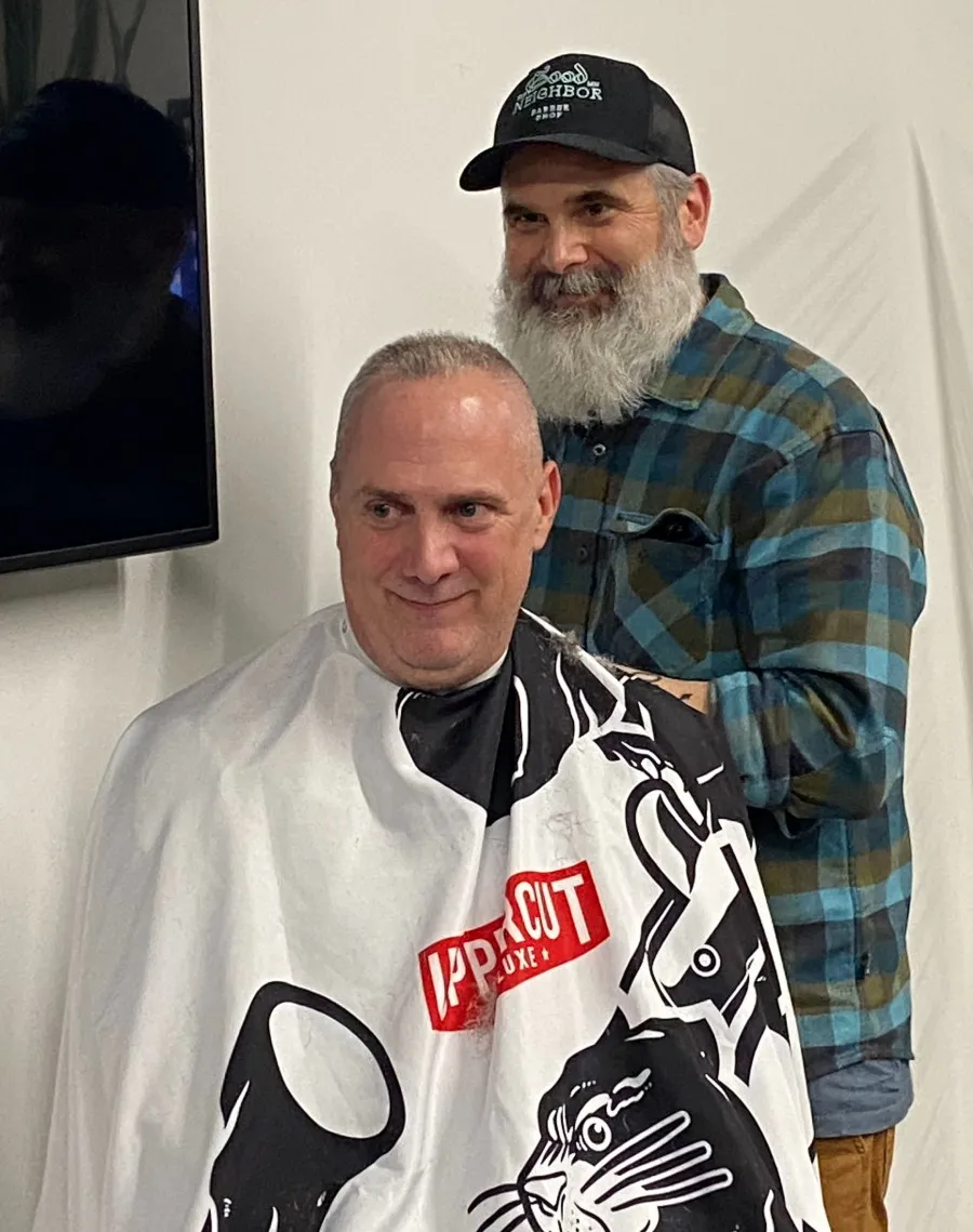Two men look off camera -- a standing man in flannel with a large gray beard, and a seated man in a barber's cape with a freshly shaved face