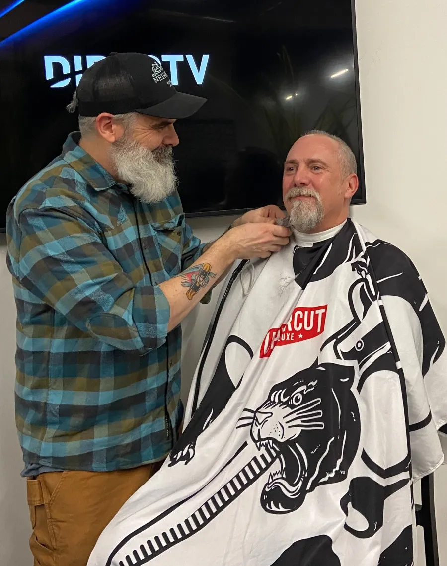 A man in a flannel shirt with a large gray beard shaves a goatee off the face of a bald seated man