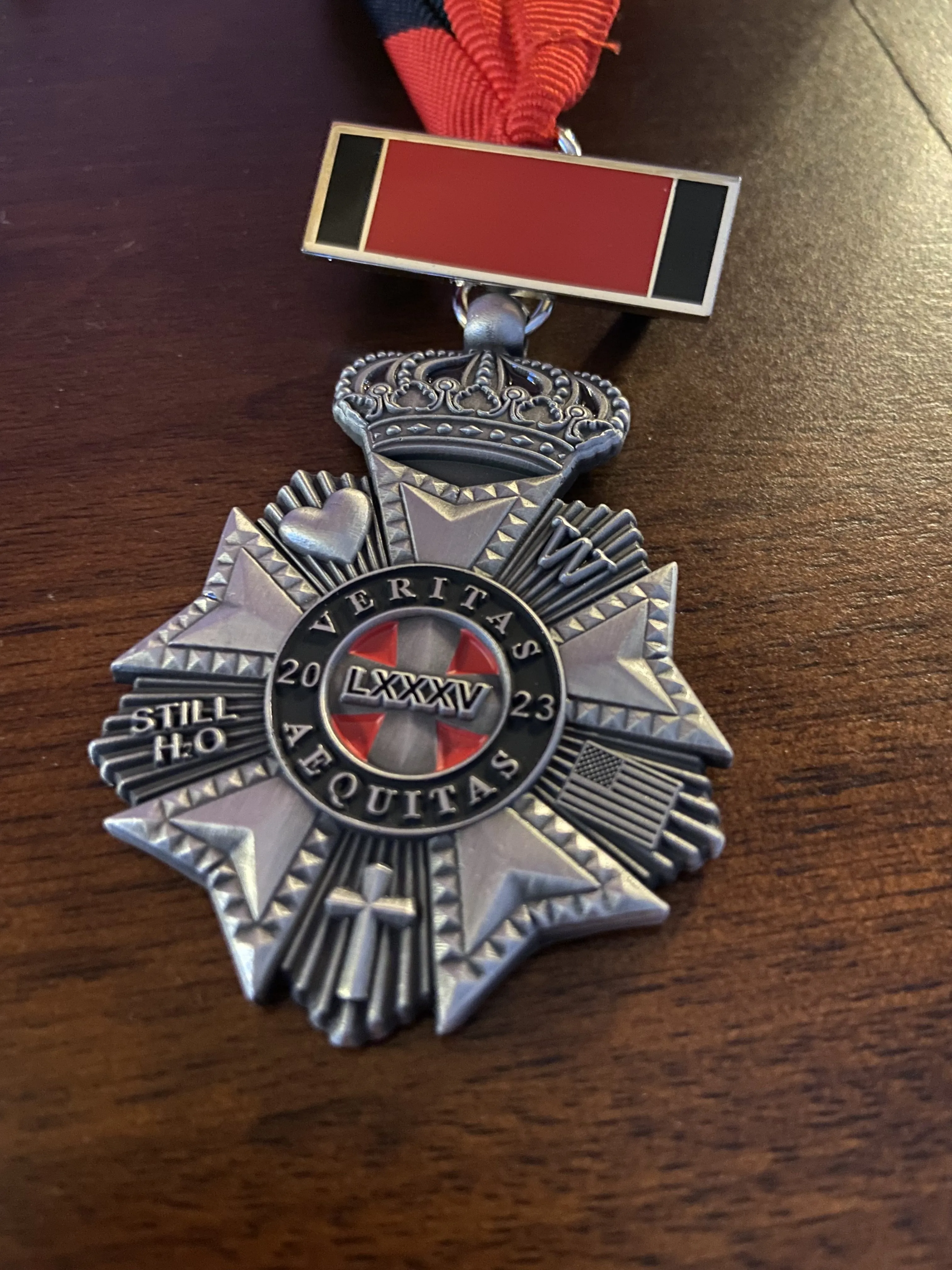 A medal sits on a table. The medal has five symbols -- heart, W, cross, American flag, "Still H2O" surrounding a center cross with the words "Veritas Aequitas"