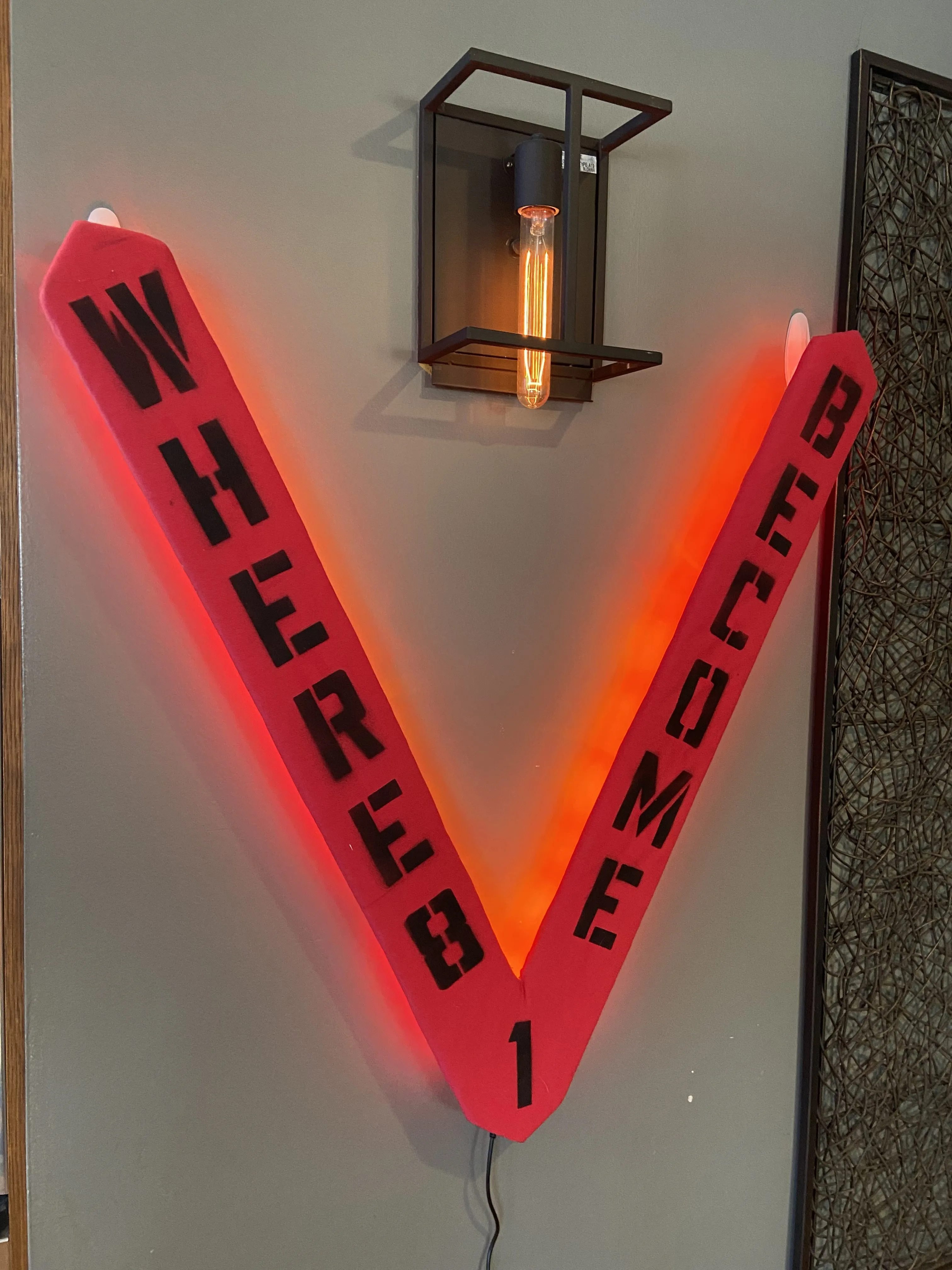 A large wooden V, painted red and lit from behind with red lights, says "Where 8 Become 1" in black stencil lettering