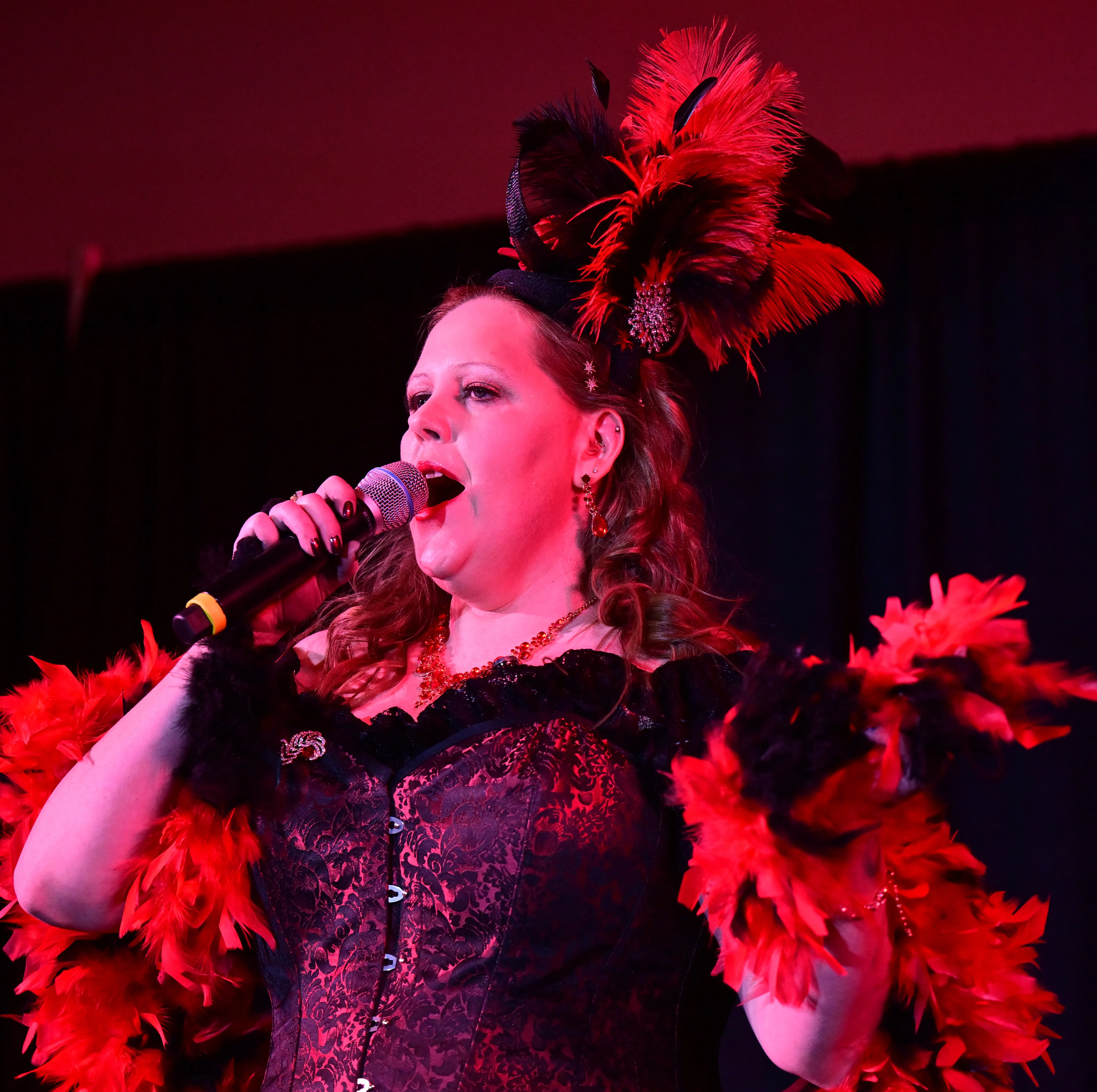 A woman in red feather boa sings into a microphone.
