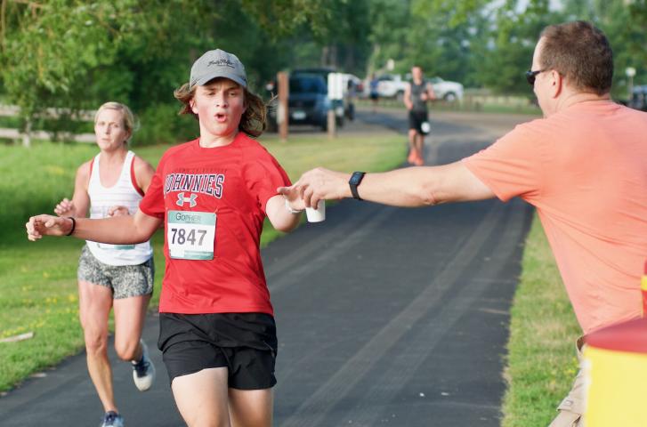 Brett Penk reaches for a glass of water offered by Jim Watt at the water station on the west side of Lake Ripley. Penk, 15, finished 10