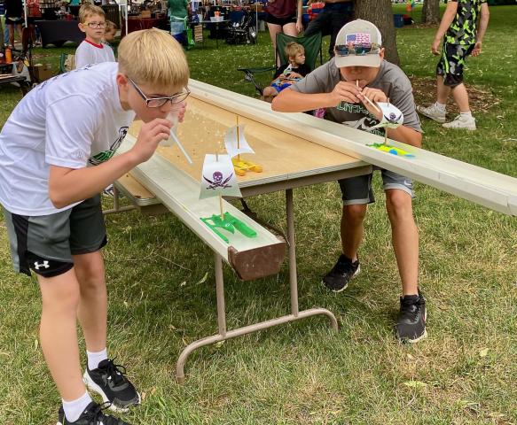 Ethan LaGow and Carter Rothstein use their own wind power — blowing through straws — to push boats down the track. The race setup was part of the Litchfield Cub Scouts booth during Art in the Park Saturday in Central Park.